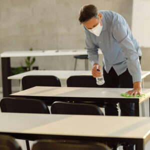 Enseignant moyen adulte portant un masque de protection du visage pendant le nettoyage des tables avec désinfectant dans la salle de classe.